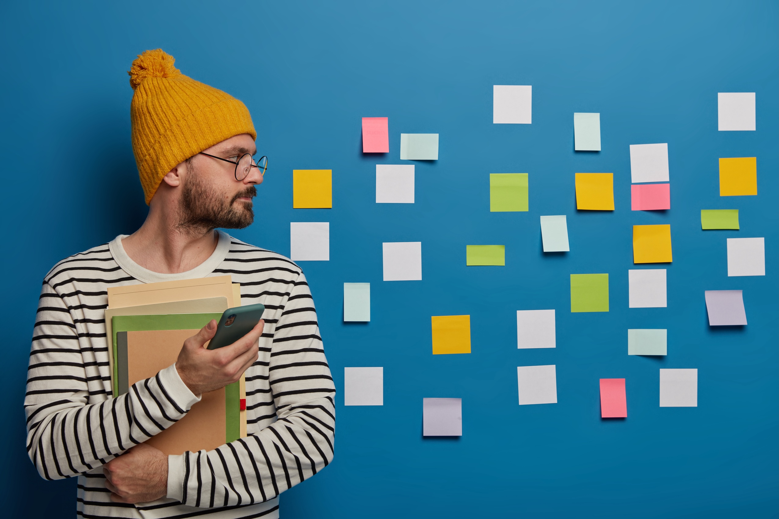 Indoor View Of Serious Bearded Man Wears Yellow Hat And Striped Jumper, Focused Aside On Wall With Sticky Notes, Uses Mobile Phone, Checks Newsfeed, Learns New Information, Holds Notepad, Books