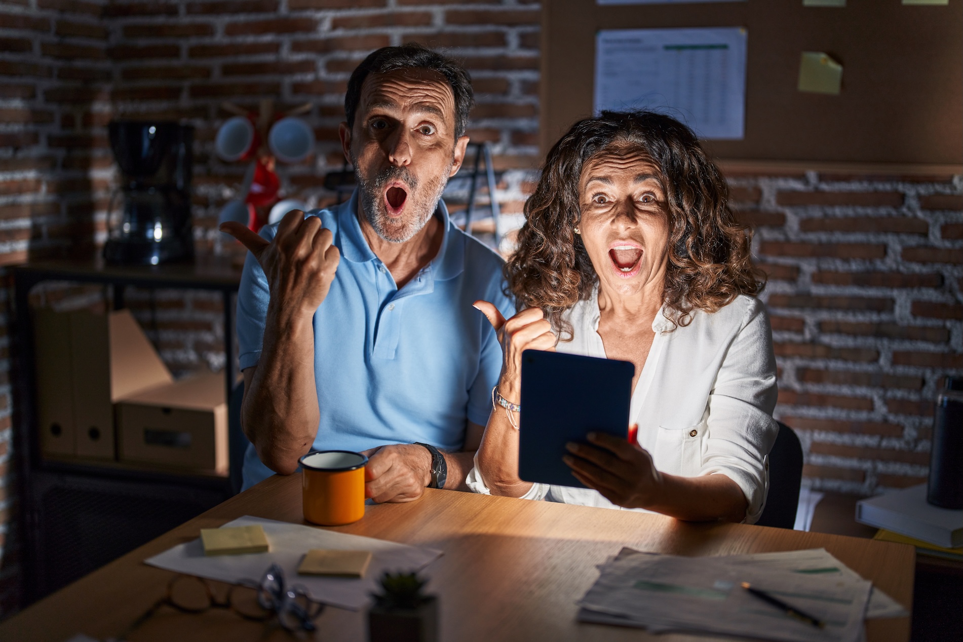 Middle Age Hispanic Couple Using Touchpad Sitting On The Table At Night Surprised Pointing With Hand Finger To The Side, Open Mouth Amazed Expression.