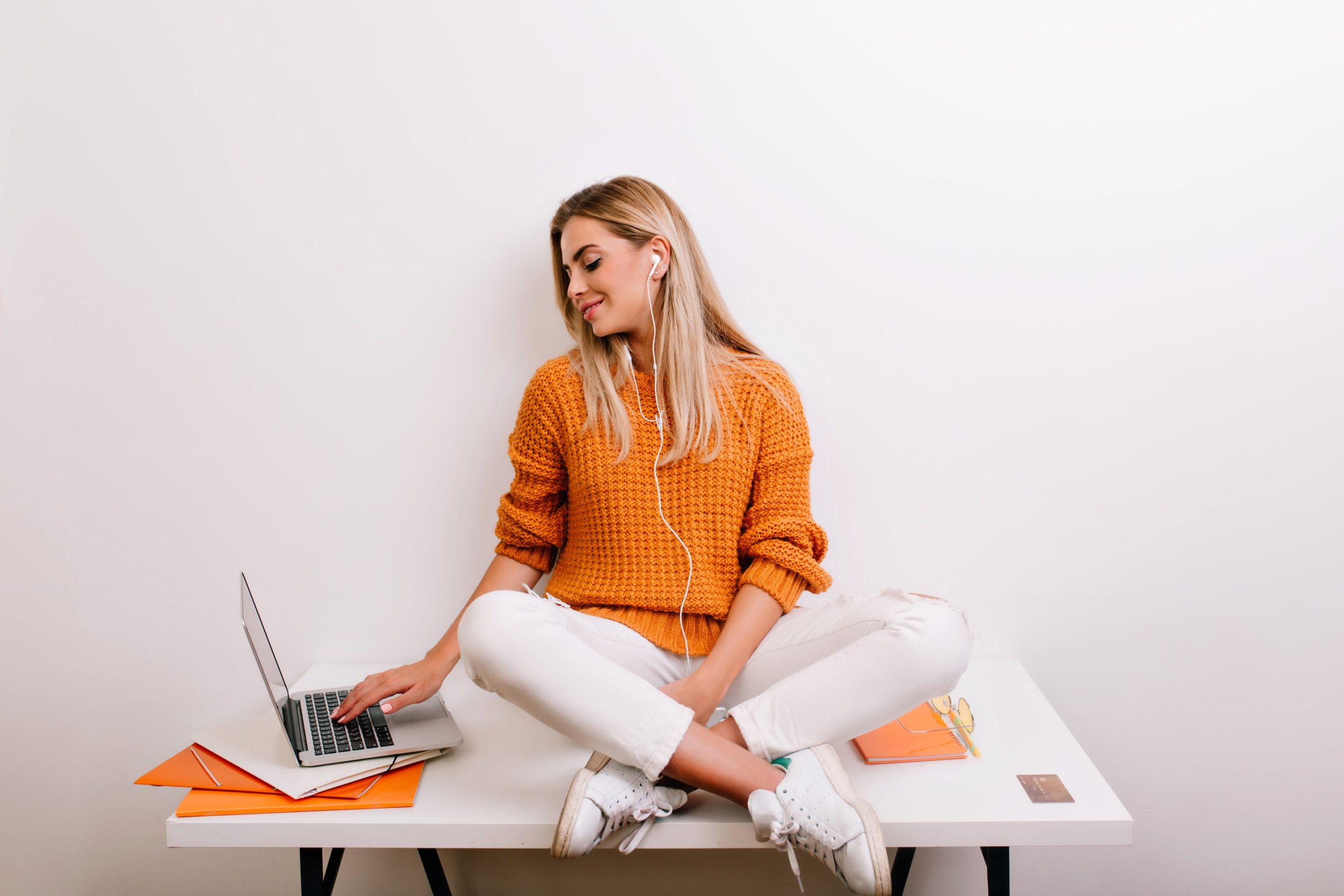 Smiling Blonde Girl Relaxing In Home, Listening Music And Typing Something On Keyboard. Indoor Portrait Of Cute Fair Haired Woman In Yellow Sweater Chilling With Laptop In Office..